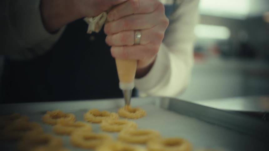 A baker using a piping bag to shape dough into circles on parchment in a bakery kitchen