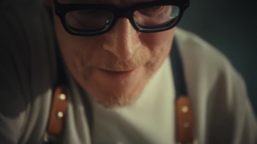 A focused male baker wearing glasses leans over a counter preparing dough in a kitchen