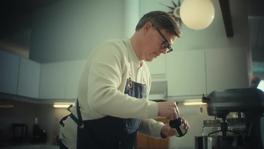 Man wearing glasses and apron adds liquid ingredient from a bottle to the mixer mixture