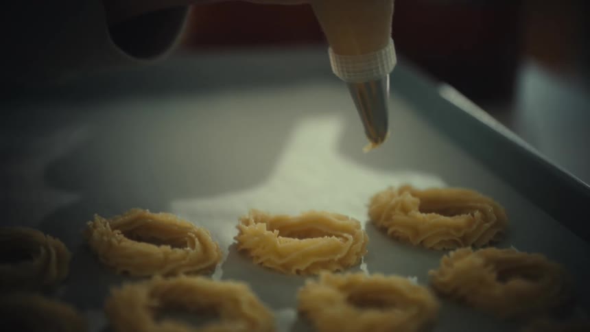 Hands use a piping bag to form dough rings on a tray