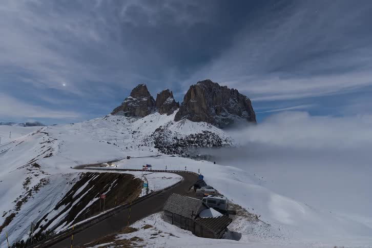 A wide night time view shows cars driving along a winding road at sella pass with light trails visible against the snow covered landscape and the jagged peaks of langkofel in the dolomites under a moonlit sky