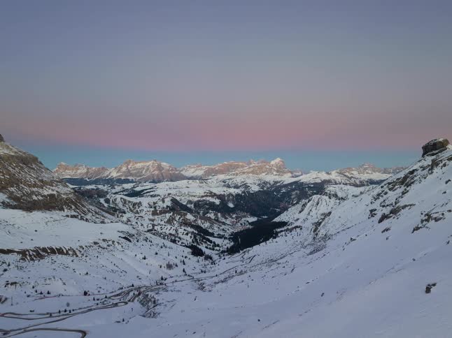 A wide aerial view shows a vast mountain range covered in snow under a pale purple and pink sky during twilight