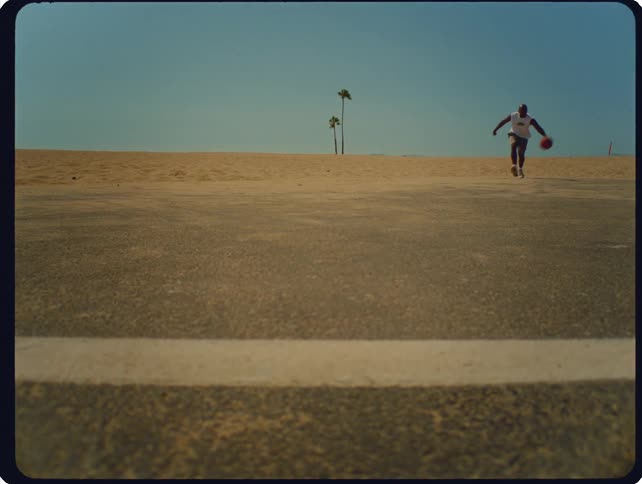 A person dribbles a basketball while running across an empty road toward a sandy horizon under a clear sky