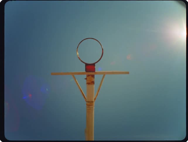 A low angle static shot shows a basketball rim without a net against a clear blue sky with a bright sun creating lens flares