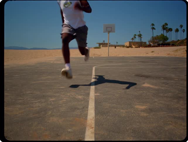 A man runs directly toward the camera across an outdoor basketball court situated on a beach under a bright blue sky