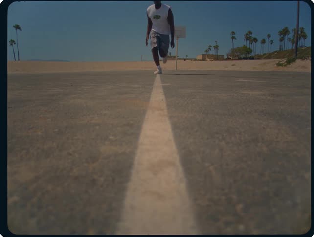 A man runs directly toward a low angle camera on an outdoor asphalt basketball court near a beach