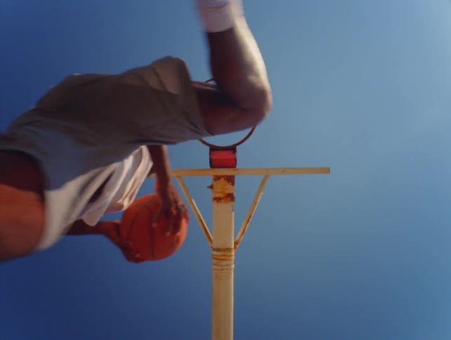 A low angle view shows a basketball player jumping over the camera to dunk a basketball into a hoop under a clear blue sky
