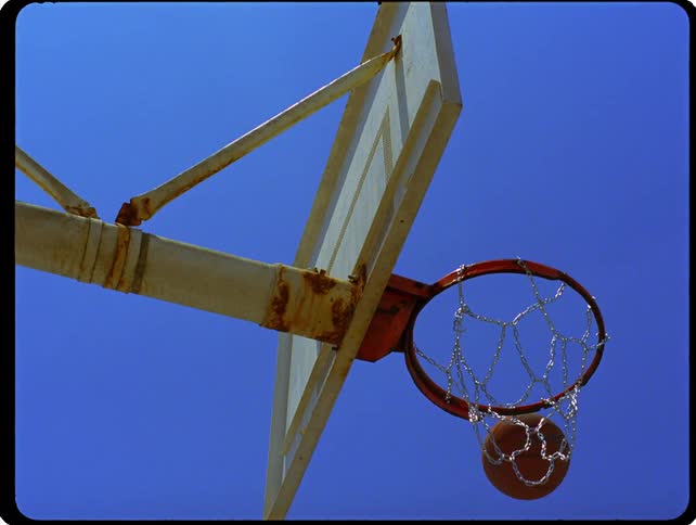A basketball passes through a metal chain hoop attached to a weathered outdoor backboard against a clear blue sky