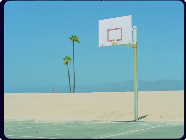 A static wide shot shows an outdoor basketball hoop standing on a court next to a sandy beach with palm trees under a clear blue sky