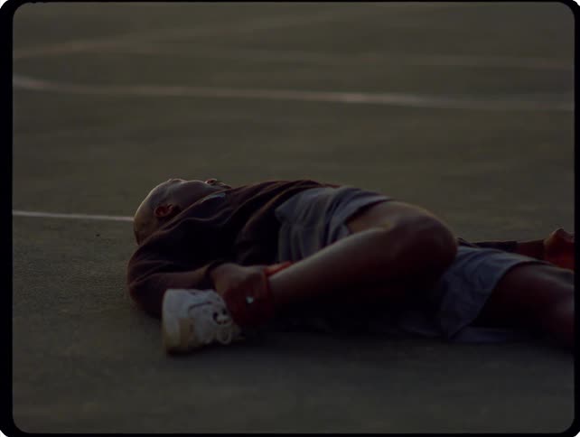 A man lies on an outdoor court and stretches his legs while resting on his back during twilight