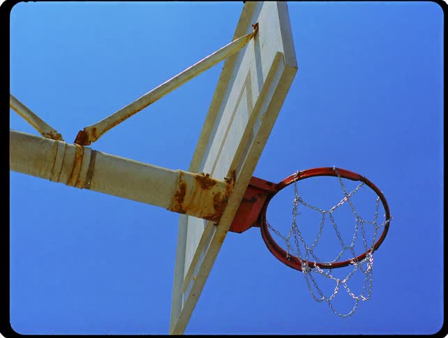 A low angle static shot shows an outdoor basketball hoop with a rusty rim and a chain net under a clear blue sky