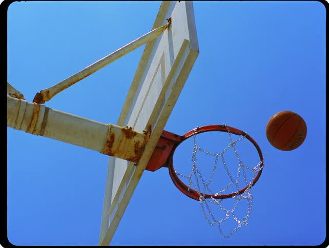 A basketball passes through a metal chain hoop attached to a weathered backboard against a clear blue sky
