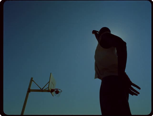 A silhouetted man takes a jump shot and makes the basket against a clear sky