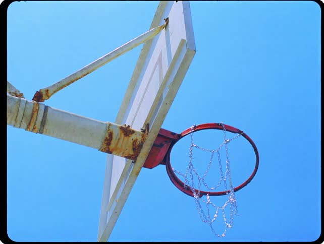 A weathered basketball hoop with a metal chain net and rusted backboard stands against a sky