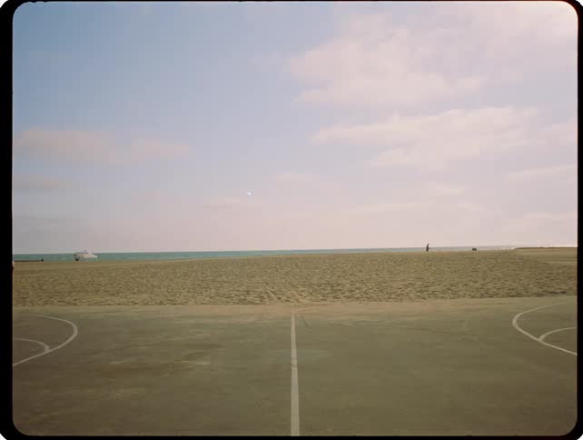 A man wearing a white tank top and grey shorts dribbles a basketball across an outdoor court located next to a sandy beach under a clear blue sky