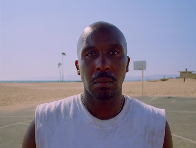 A close static shot shows a bald man with a serious expression looking directly into the camera while standing outside on a sunny day