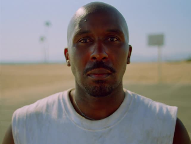A portrait shot of an athletic black man looking directly at the camera on an outdoor basketball court