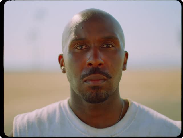 Man with shaved head and short beard looks forward in an outdoor setting