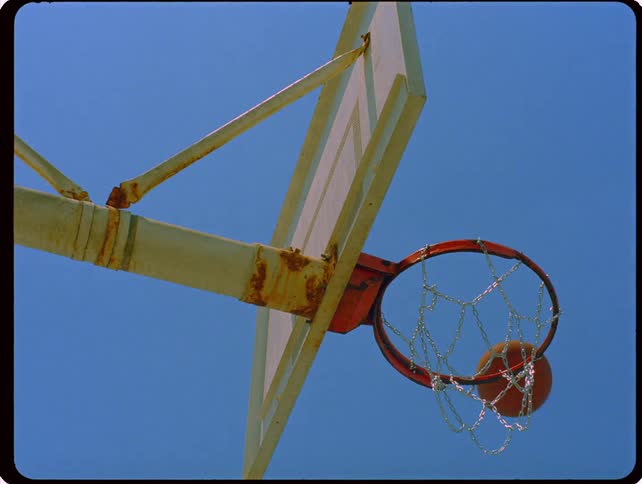 A basketball passes through a metal chain hoop mounted on an outdoor backboard against a clear blue sky