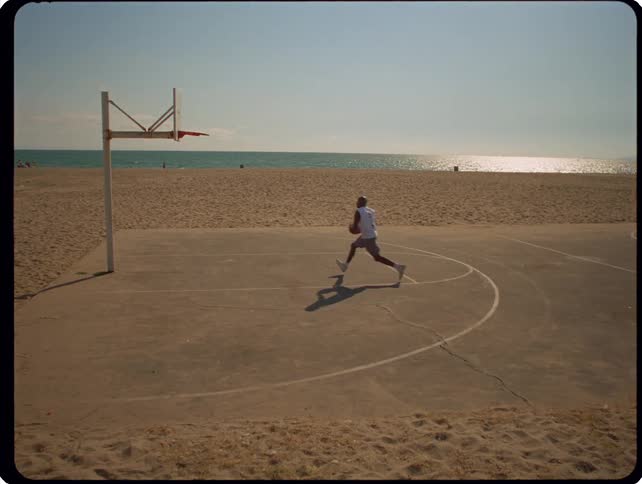 A man dribbles a basketball across an outdoor court located on a beach and performs a layup while the sun reflects off the ocean water
