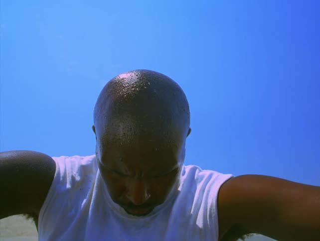 A young man sitting in a white tshirt sweating and looking tired as he bows his head to the ground under the sun