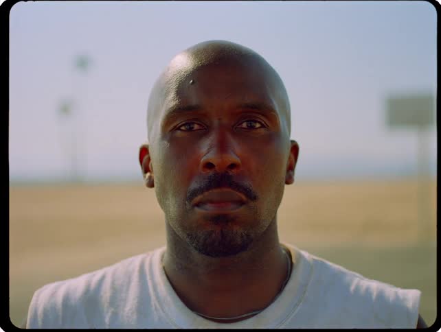 A close static shot shows a man with a shaved head and a serious expression looking directly into the camera while standing outdoors on a bright day