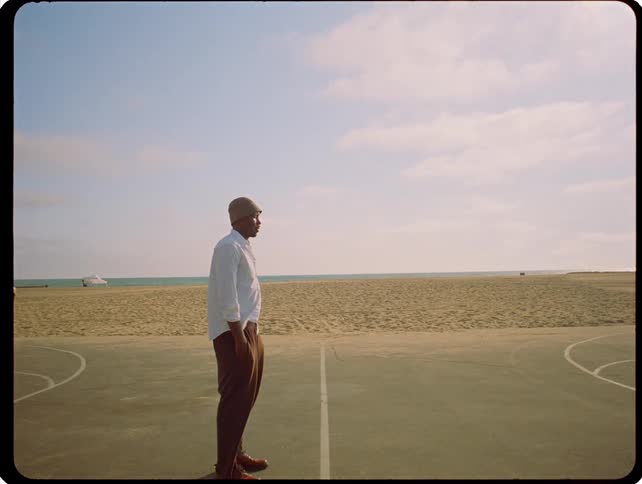 A man stands on a seaside basketball court looking toward the horizon as a sudden bright white flash illuminates the frame before returning to the calm beach scene