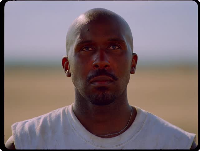 Man with a shaved head and a sleeveless shirt looks ahead in an outdoor setting