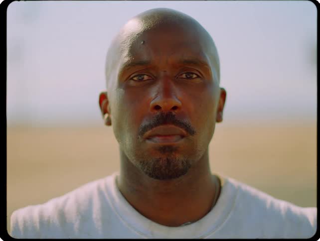 A close static shot shows a man with a shaved head and a serious expression looking directly into the camera while standing outdoors on a bright day