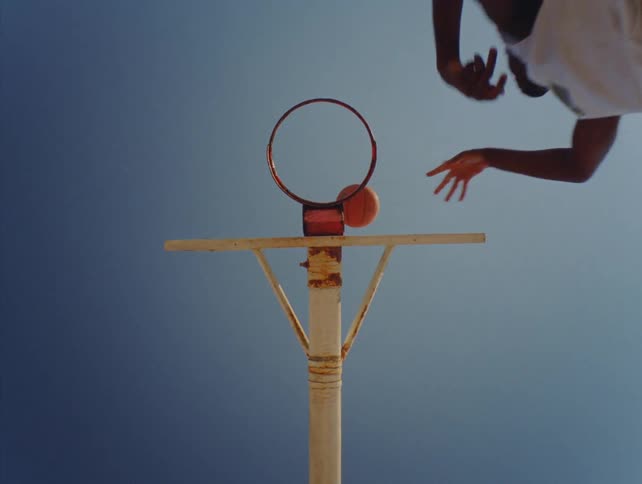 From a low angle a basketball player leaps into the frame to attempt a layup against an outdoor rim the orange ball soaring upward and missing the hoop against a hazy bright sky