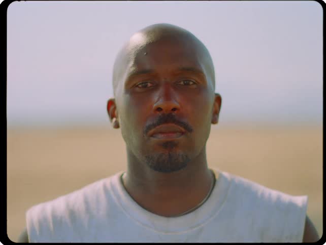 A man maintains a steady unflinching gaze toward the camera before a sudden bright light flash washes over the scene momentarily illuminating his features against the hazy outdoor background