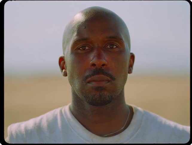 A close static shot shows a man with a shaved head and a serious expression looking directly into the camera while standing outdoors on a bright day