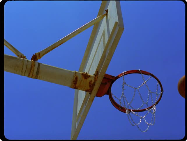 A basketball hovering above a hoop about to fall to the ground at midday