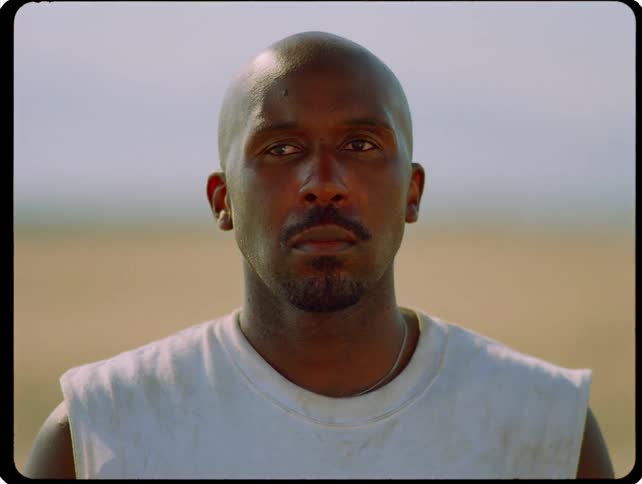 A man stands still and looks directly forward with a steady gaze his expression calm and composed against a blurred sundrenched outdoor landscape