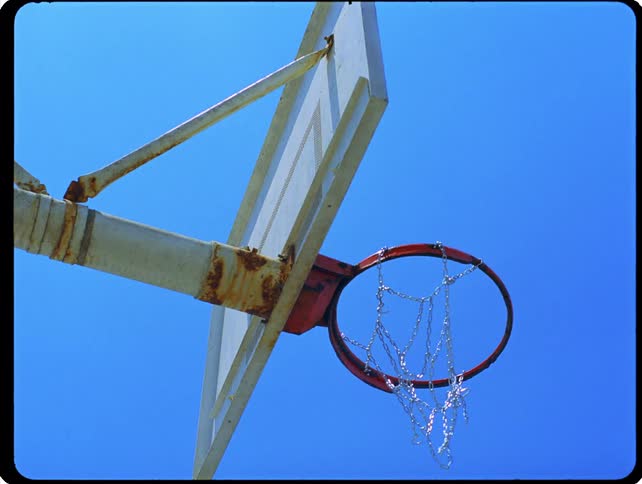 Against a clear blue sky a basketball flies through the air and rattles through a metal chain net on a weathered outdoor hoop reflecting a classic afternoon of streetball and recreation