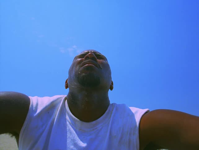A sweaty man in a white tank top looks up toward the sky and then back down against a clear blue background