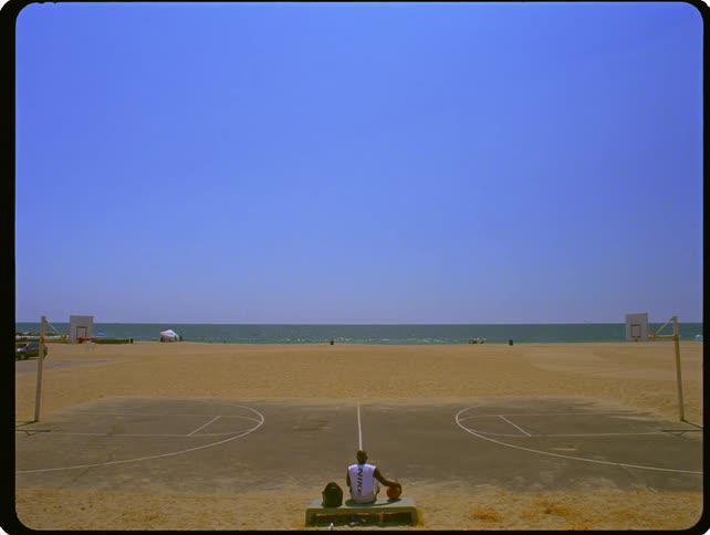 A man sits alone on a bench at a beachside basketball court looking out at the ocean