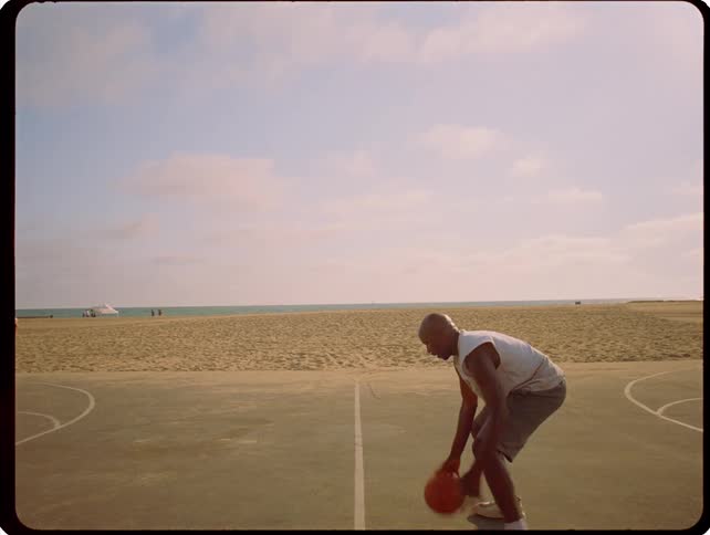 A man dribbles a basketball on an outdoor court near a beach