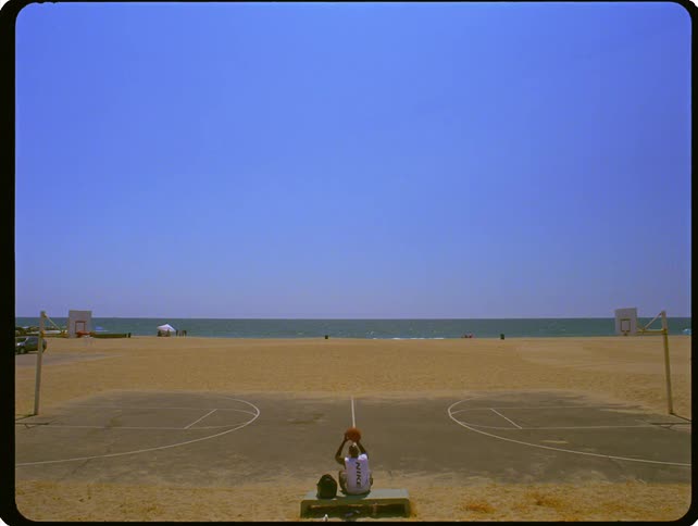 A lone basketball player sits on a bench facing a court on a beach and throws a basketball into the air
