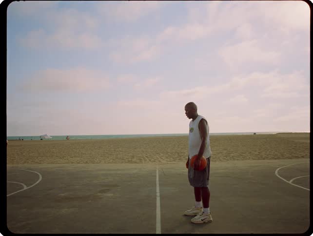 A man stands alone on an outdoor basketball court at the beach while holding a basketball under a cloudy sky