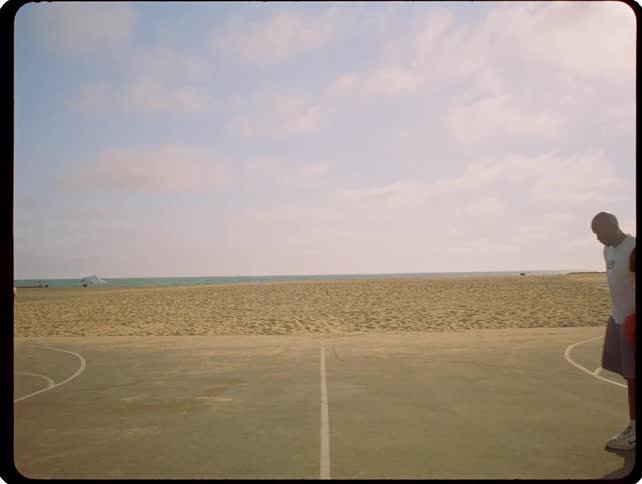 Two men play basketball on an outdoor court situated next to a beach