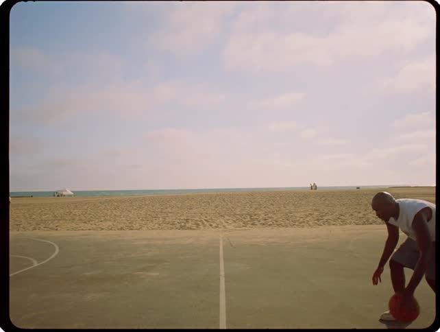 A man dribbles a basketball in a slow and controlled manner on an outdoor court at the beach under a sky filled with soft clouds
