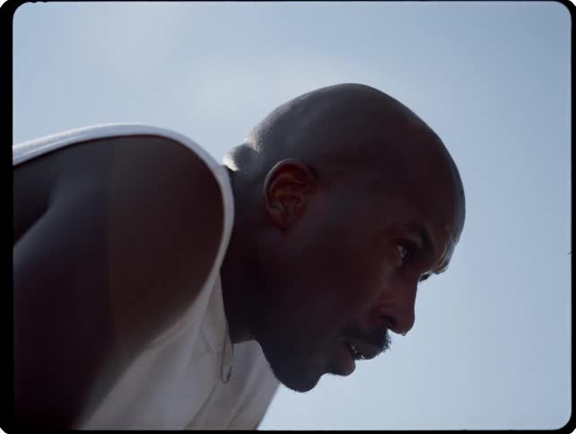 A man with a shaved head and a mustache looks down while breathing heavily after a workout on a beach basketball court