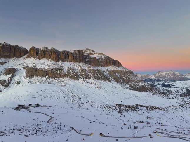An aerial shot shows a massive snow covered mountain range during sunset as the orange light fades and a moon appears in the pink sky