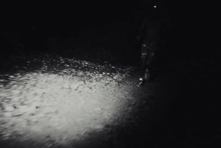 A shirtless man wearing shorts runs on a dark dirt path at night while a bright light illuminates the ground and his legs as he moves through the shadows