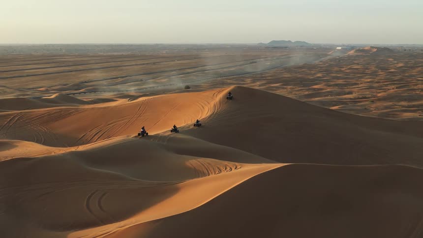 A group of people rides quad bikes over large orange sand dunes in a desert landscape during a clear day as white vehicles park in the distance