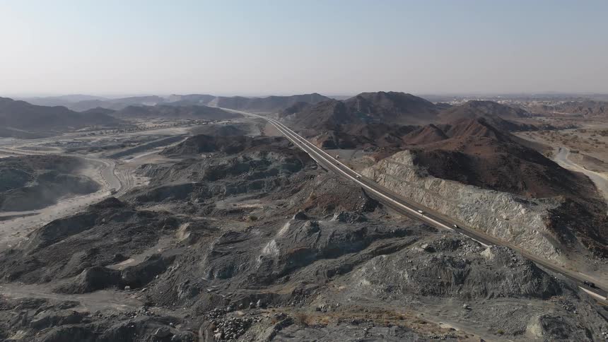 An aerial view shows a long paved highway stretching through a vast rocky desert landscape with dark mountains and industrial excavation areas under a clear sky