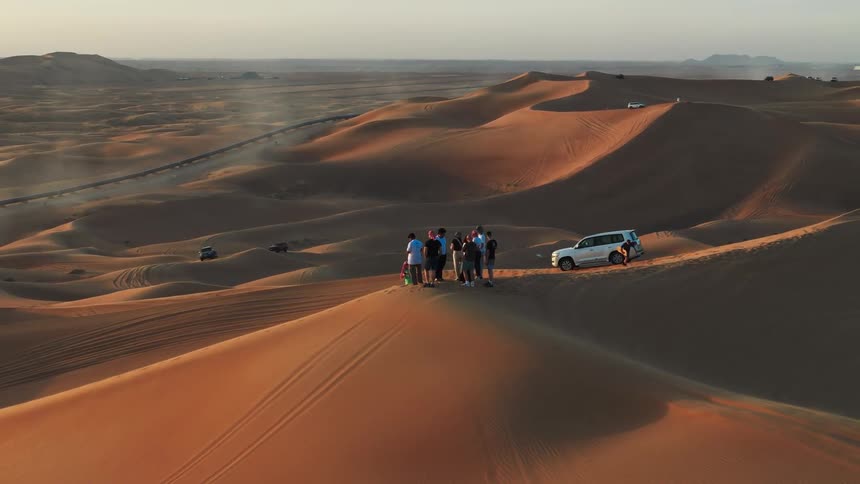 Group of people on top of a sand dune with a road and vehicle visible in the desert background