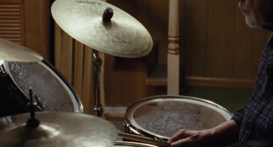 An elderly man picks up drum sticks and plays the drums