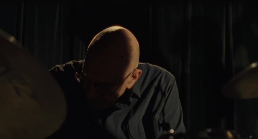 Bald man wearing glasses seated between drum cymbals in a dark setting